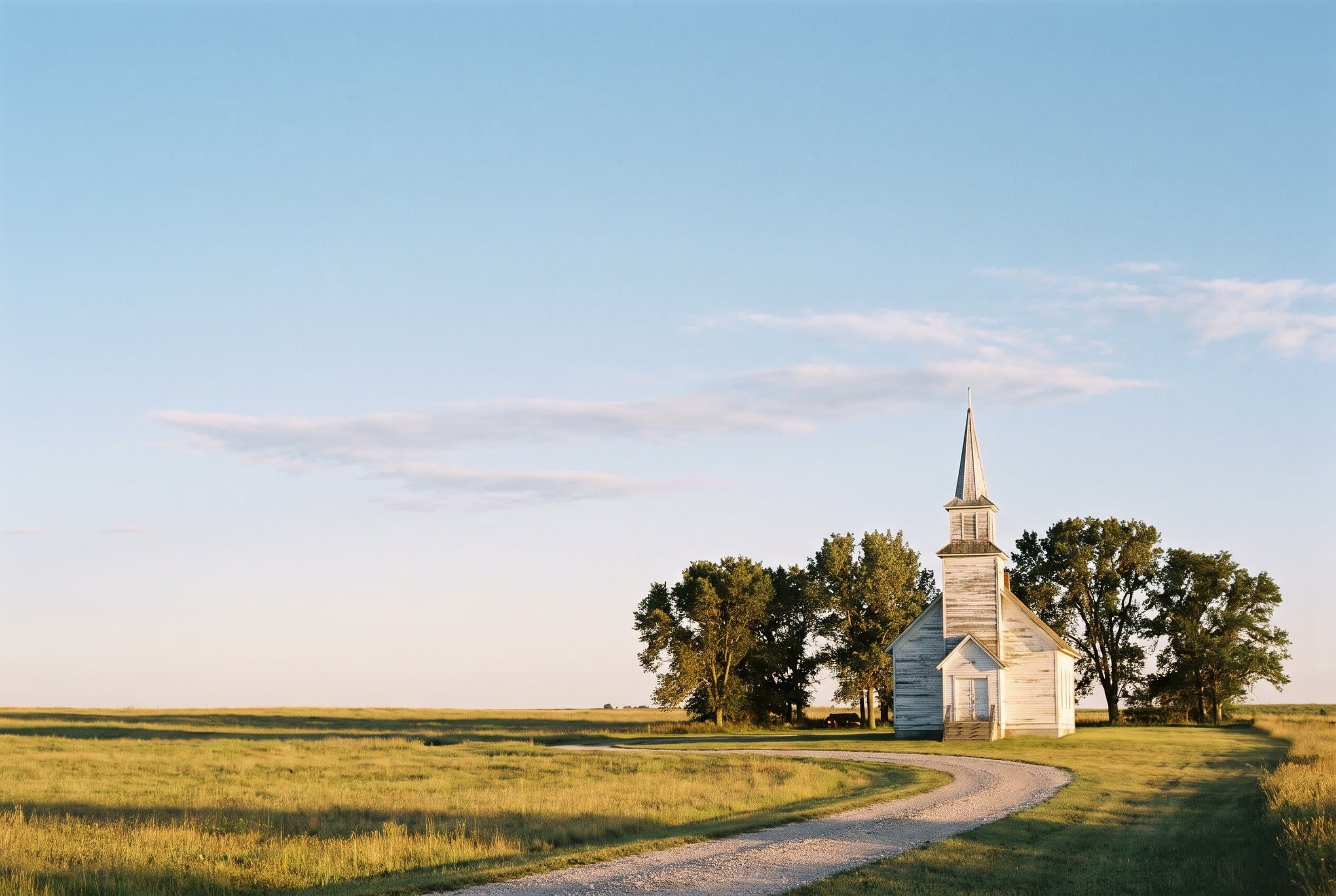 A weathered white wooden church with a tall steeple at the end of a gravel drive in open country, under a wide late-afternoon sky