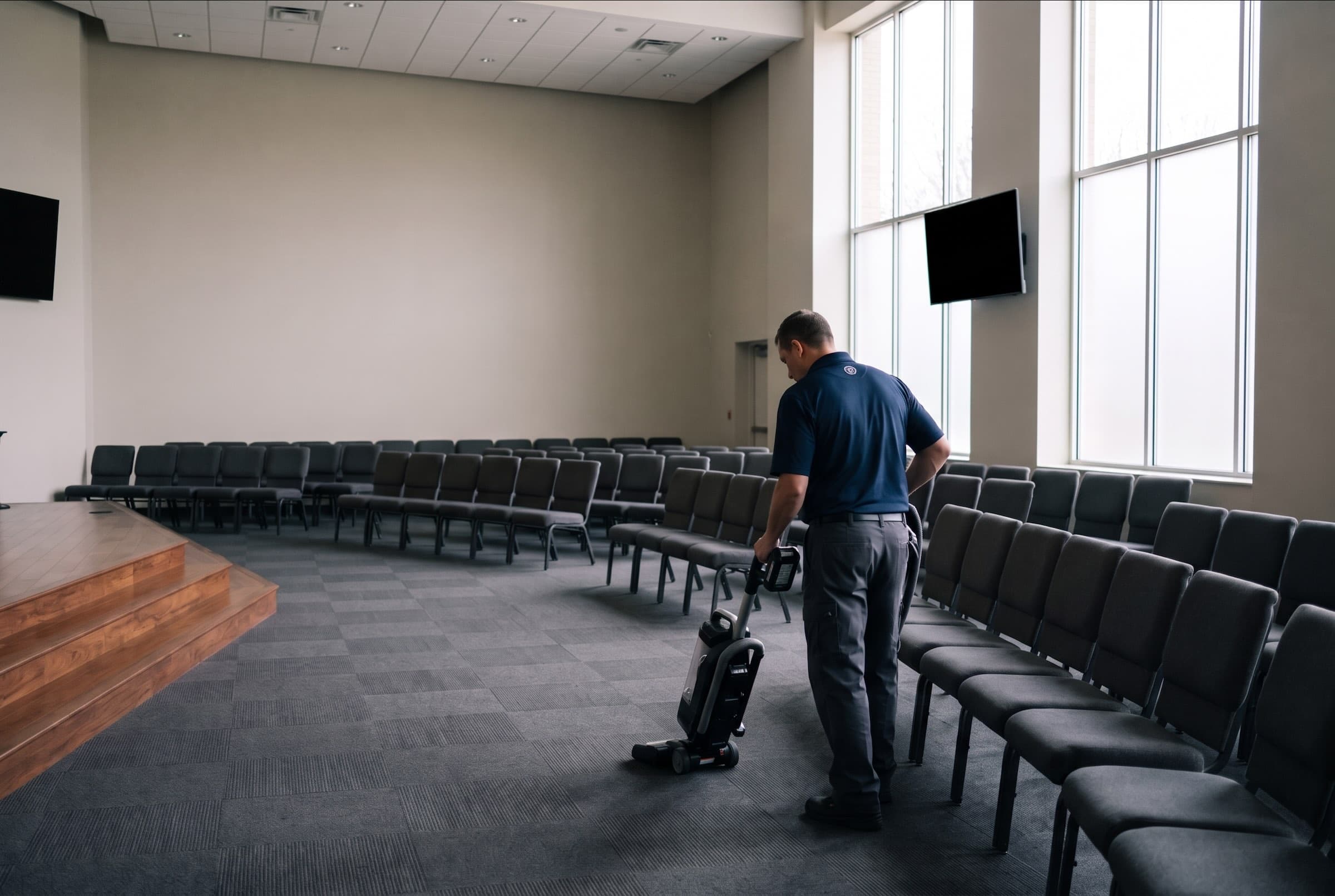 A custodian in a navy work polo vacuuming the carpet tile aisle of a modern church auditorium with a cordless vacuum, between rows of gray upholstered chairs