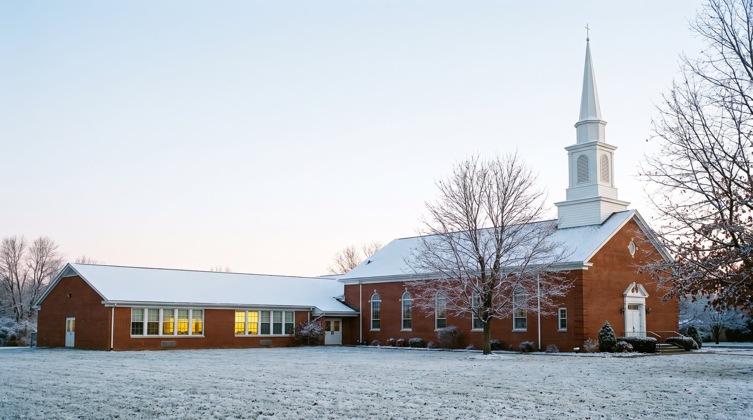 A red-brick church with a white steeple and a one-story school wing on a frosty early-winter morning, lights on in the classroom windows