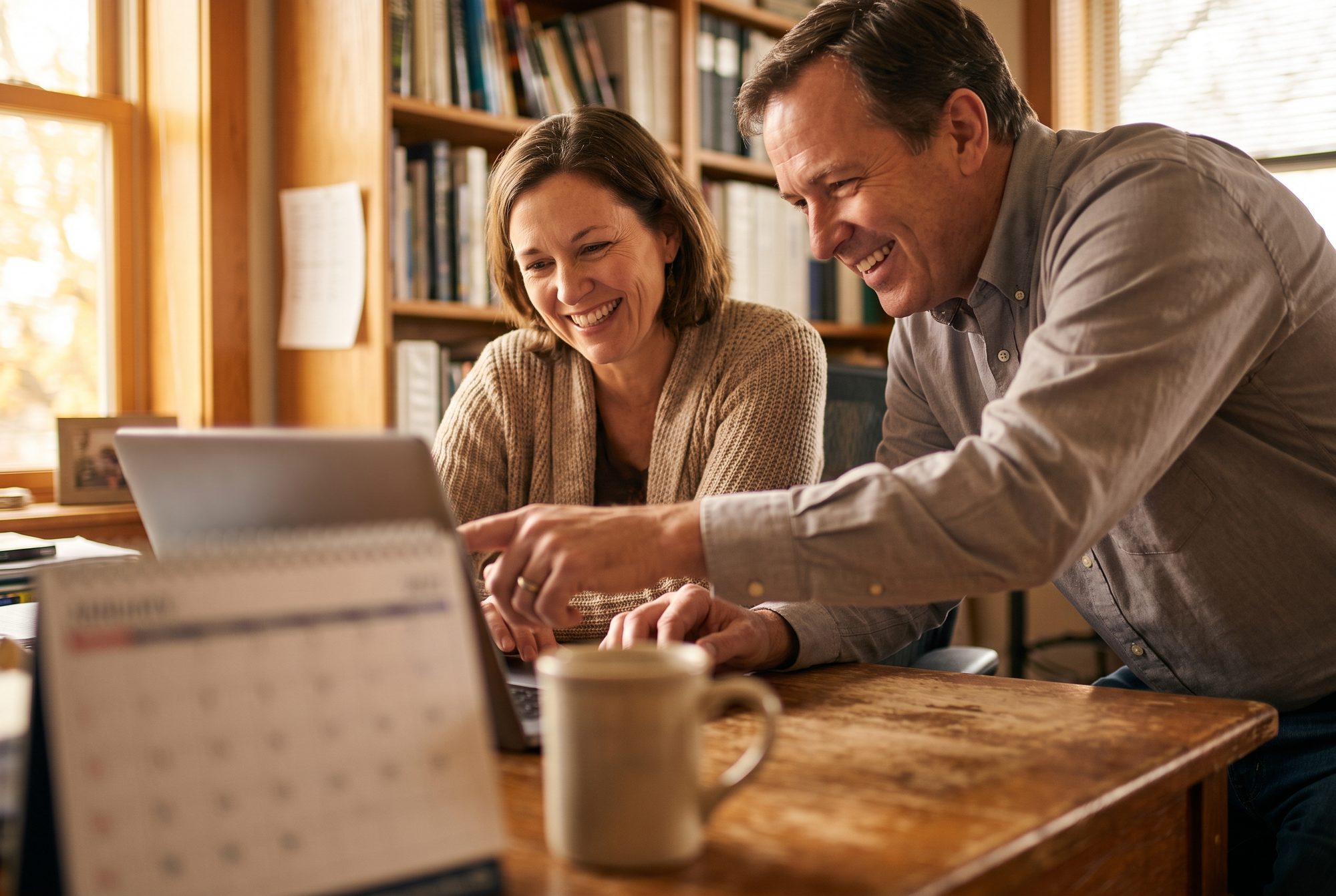 A church administrator and a school principal laugh together as they plan the year on a laptop at a wooden table, a desk calendar and a coffee mug in the foreground