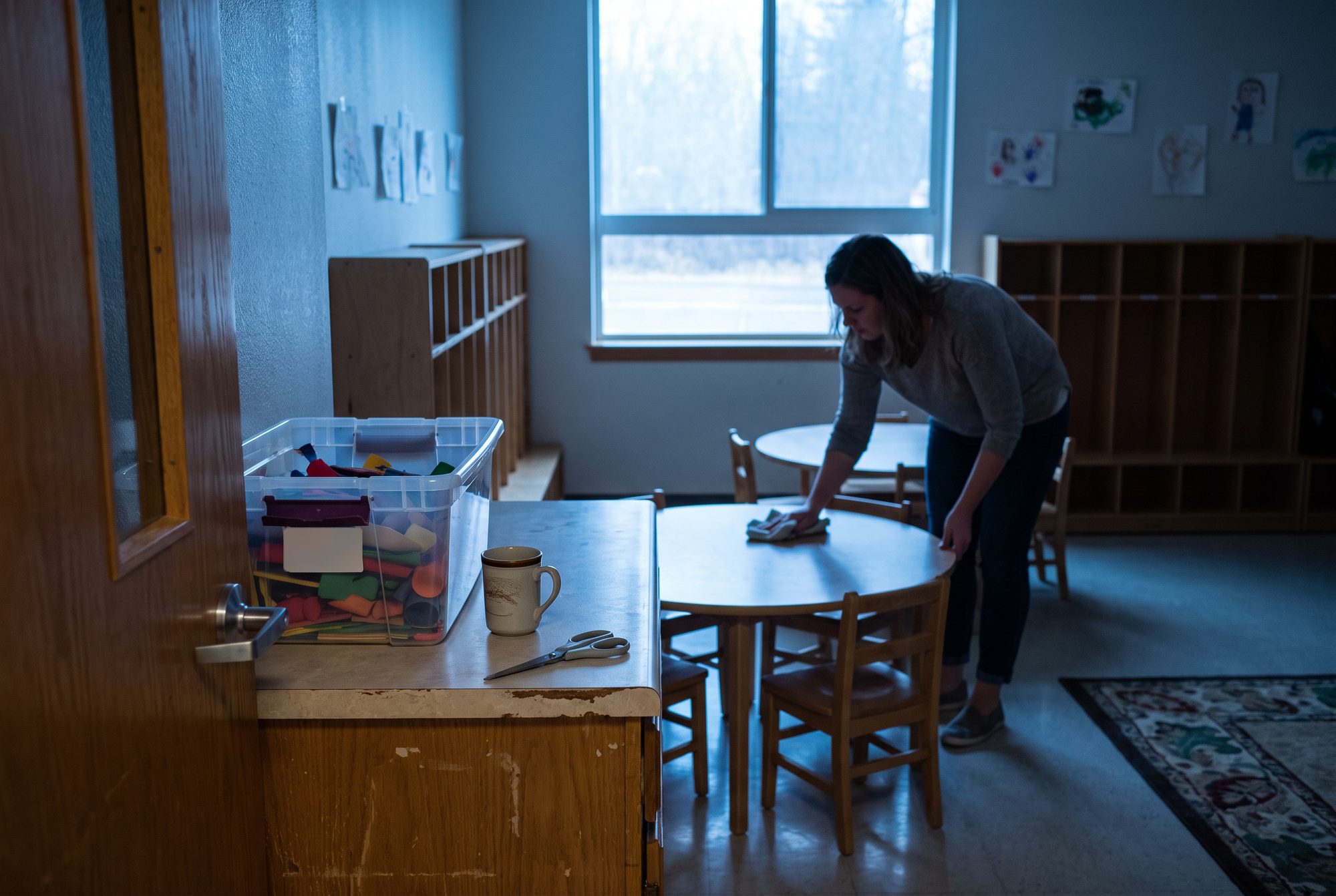 A preschool teacher wipes down a low table early on a Monday morning while a tote of Sunday school craft supplies, a coffee mug and a pair of adult scissors wait on the counter by the door