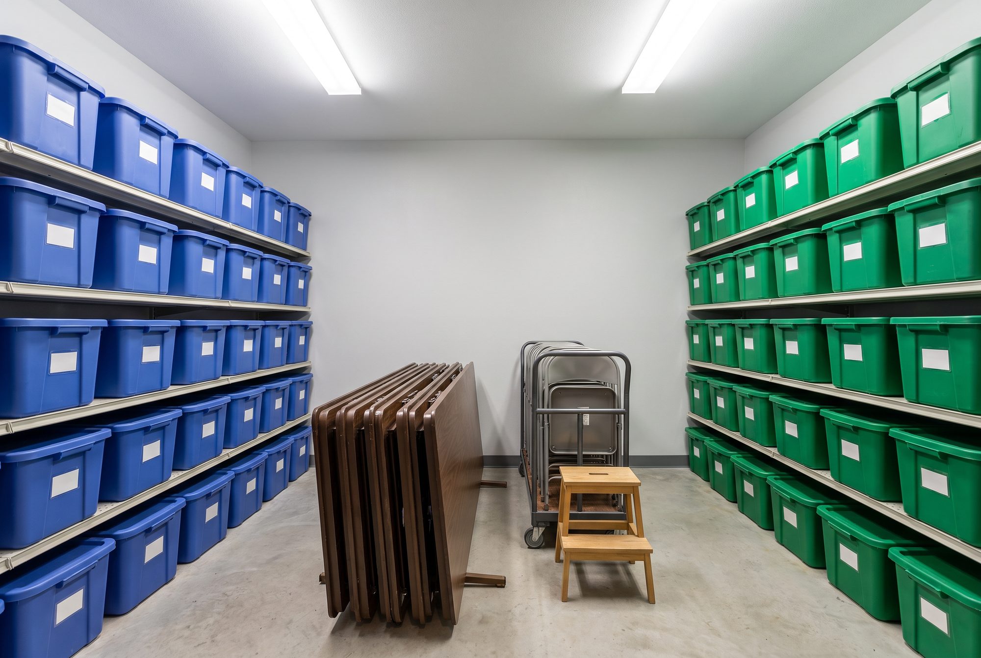 A storage closet with blue bins on the shelves along one wall and green bins along the other, folding tables, a chair cart and a step stool in the middle